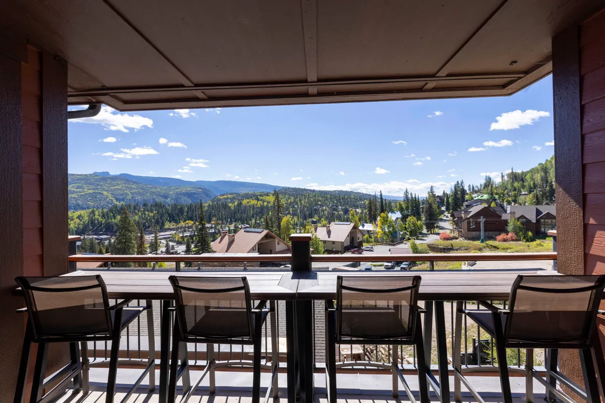 This image displays a serene outdoor seating area with a table centerpiece, overlooking a sweeping mountain view. People are seen relaxing with drinks and socializing, with the tranquil backdrop of the mountains, suggesting an ideal spot to unwind after a productive day.