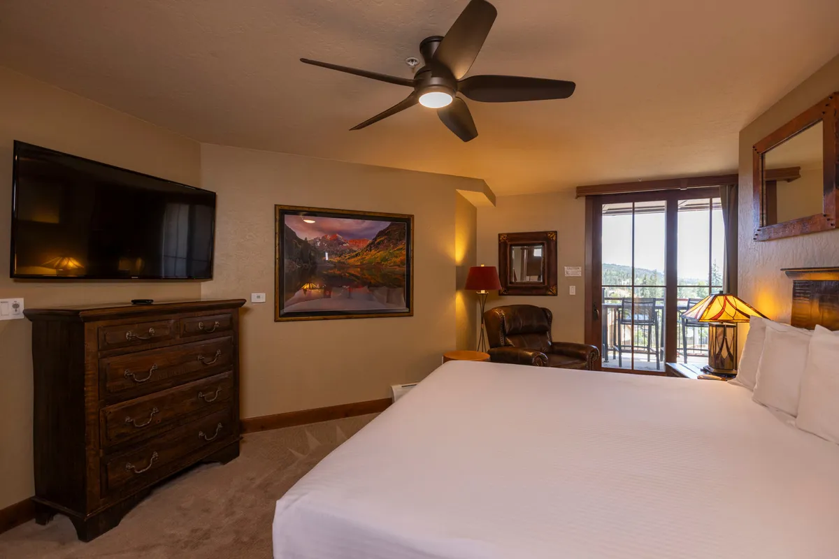 A serene bedroom featuring a comfortable bed with plush bedding, a large flat-screen television set atop a chest of drawers, and balcony doors that let in natural light. Tasteful artwork adds to the calming, personal feel of the space.