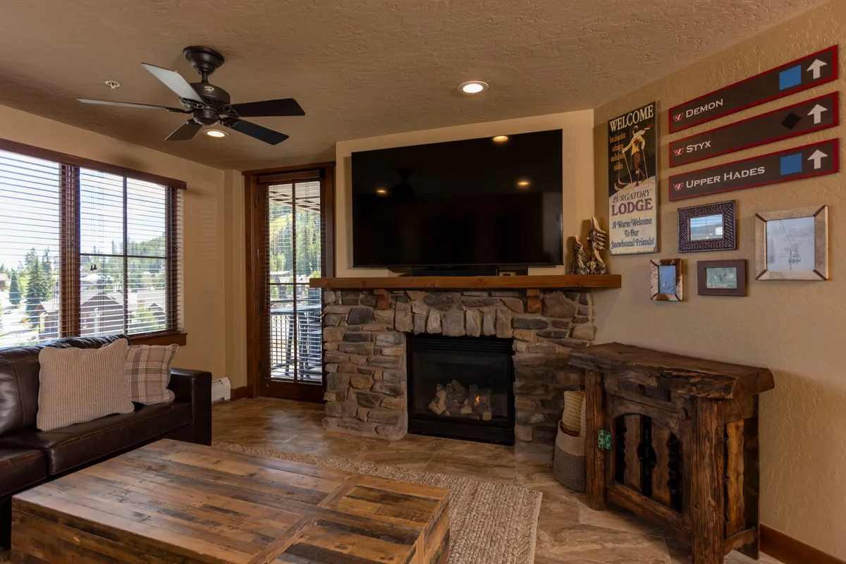 A bright and inviting living space featuring a comfortable couch, a robust stone fireplace for post-ski relaxation, a large TV for streaming or game nights, and a wooden coffee table that can easily double as a board game platform. The window view adds a touch of mountain ambiance.