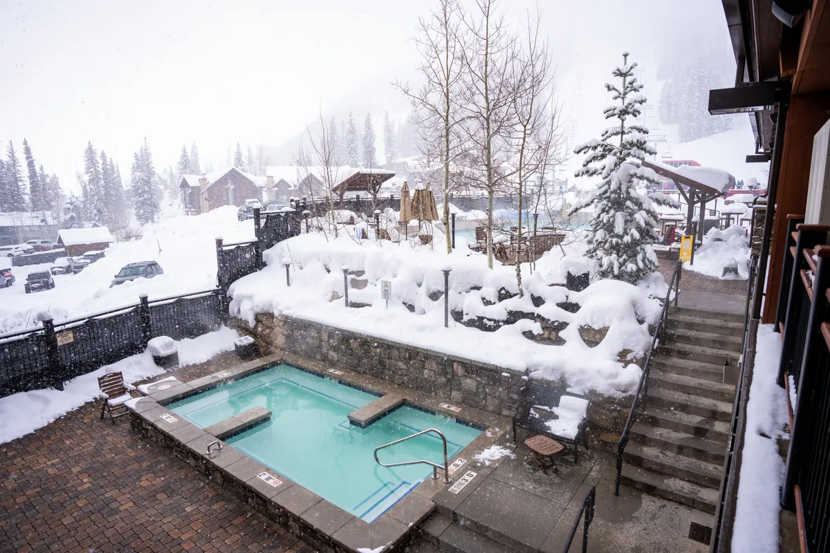 A steaming outdoor hot tub sits amid a magical snow-covered landscape with frosted trees and stonework, evoking serenity and luxury. Steps lead down to the inviting waters—perfect for post-ski relaxation and socializing in the crisp mountain air.
