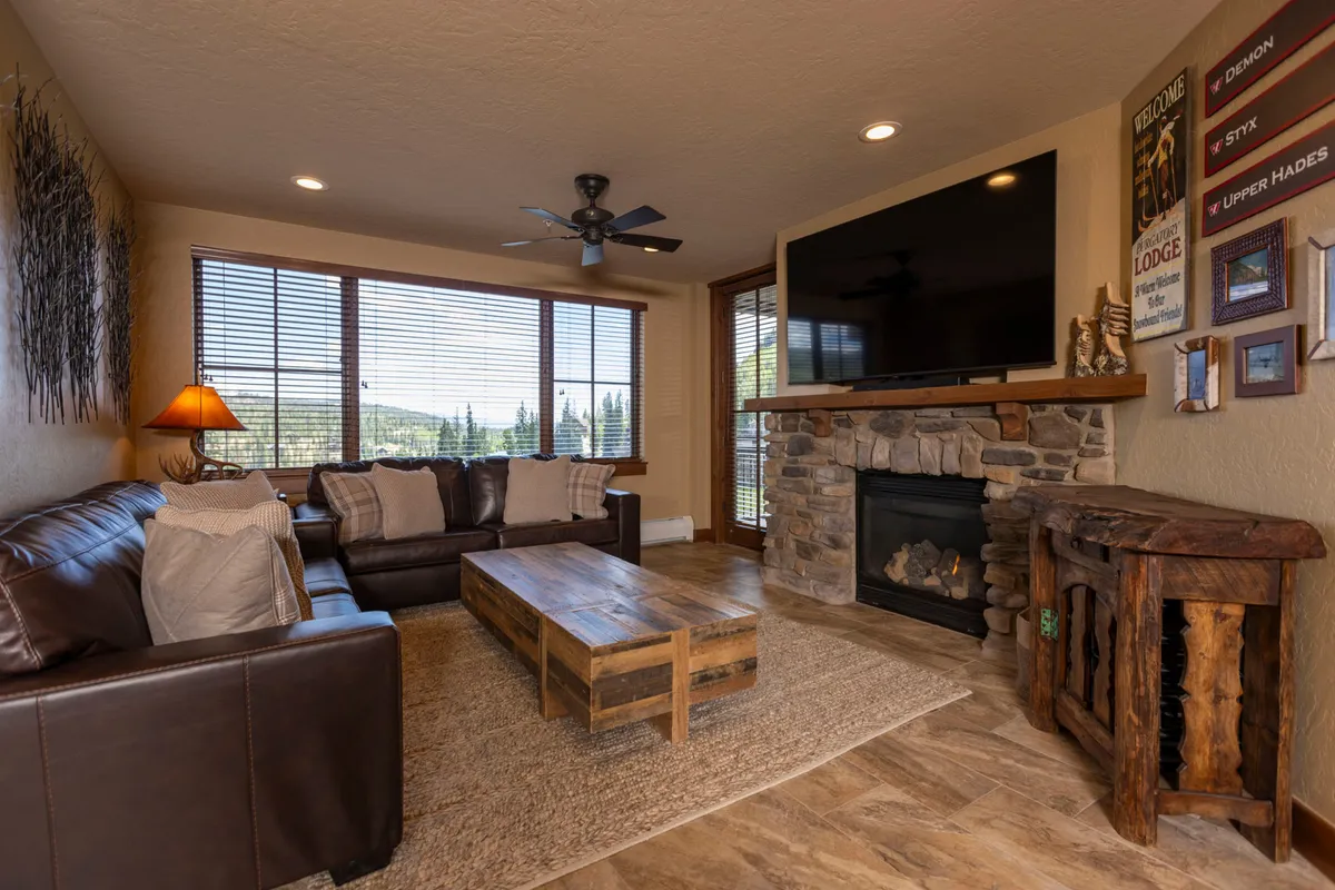 The image showcases a warm and inviting living room featuring a plush leather sofa, glowing fireplace, wooden coffee table set for games or gatherings, and expansive windows framing mountain sunsets. The space exudes comfort and relaxation, ideal for unwinding after a day on the slopes.