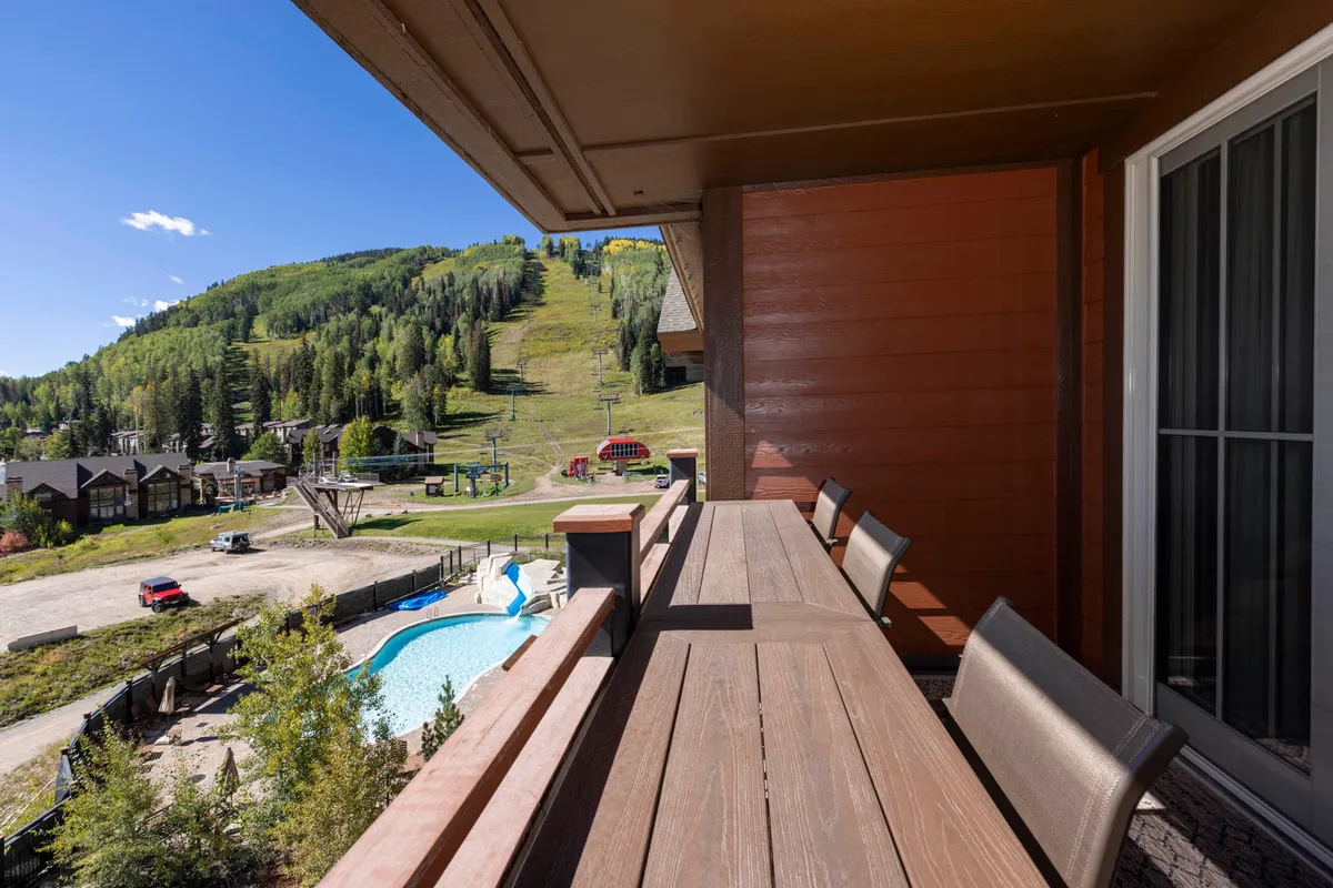 A spacious balcony overlooks a sparkling swimming pool, with the majestic mountain range and ski lift clearly visible in the background. Comfortable seating and lush greenery invite relaxation, while the vibrant pool area hints at both leisurely sunbathing and social gatherings.