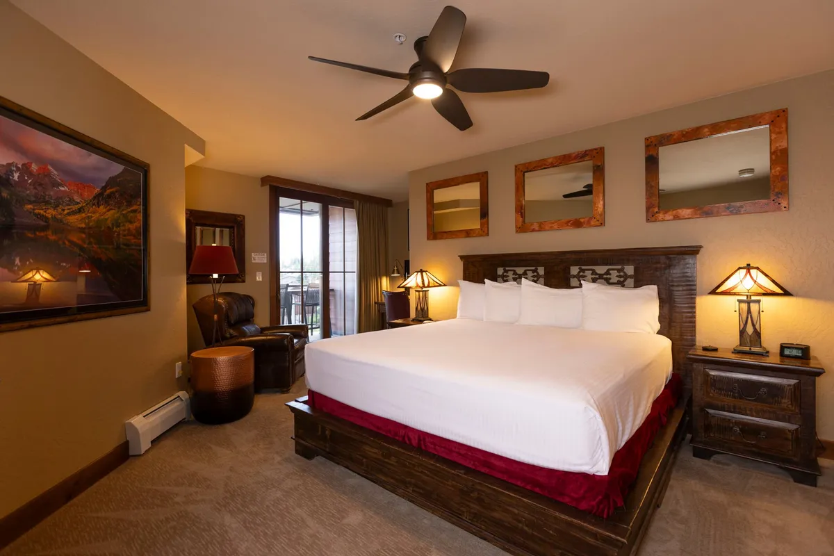 A king-size bed with soft, inviting linens dominates the room, flanked by stylish decorative mirrors and warmly lit lamps. There's an armchair in the corner perfect for reading, and a landscape painting above the bed adds a tranquil touch. Natural light filters in, creating a soothing and restful atmosphere.