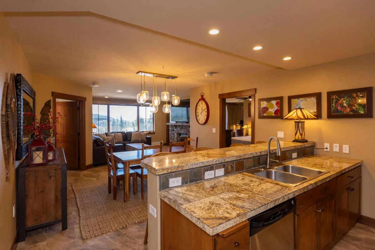 A wide-angle view showcasing the open kitchen, dining area, and living space, framed by large windows that let in abundant natural light. The kitchen island facilitates group cooking, while the dining table invites shared meals and conversation. Artful wall decor and cozy furnishing further enhance the sense of comfort.