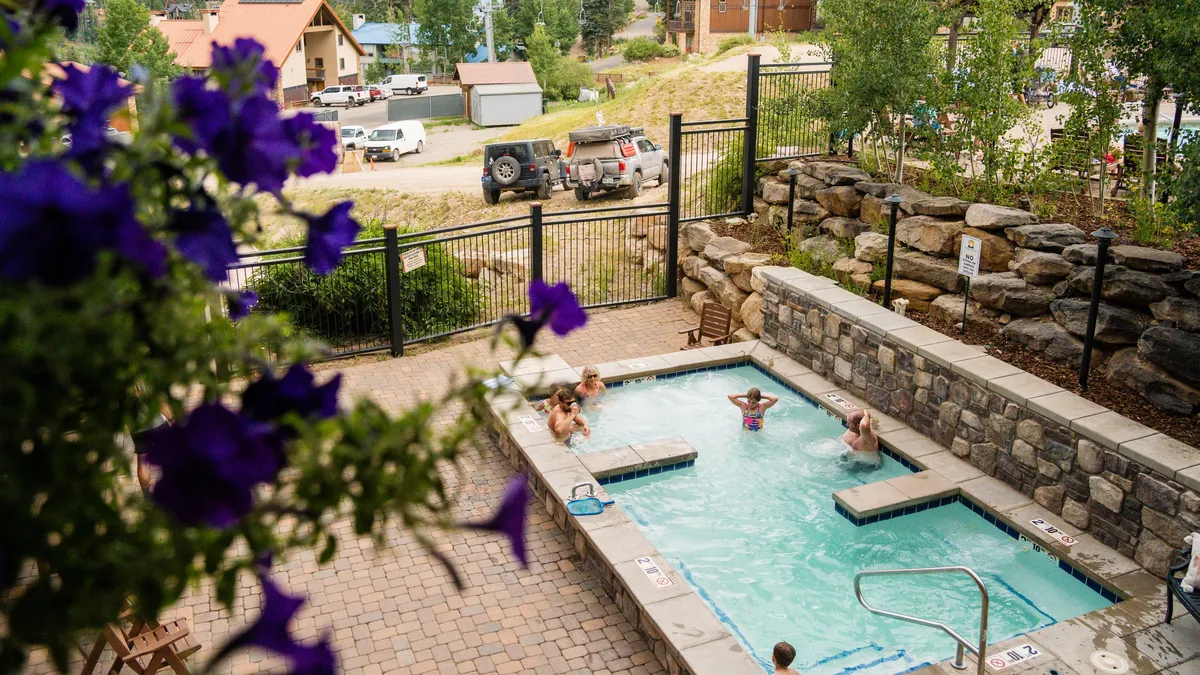 An inviting outdoor pool area framed by flowering plants and lush greenery, featuring a relaxing hot tub and plenty of space for lounging. The setting is tranquil, suggesting both family fun and peaceful relaxation in the sun or under the stars.