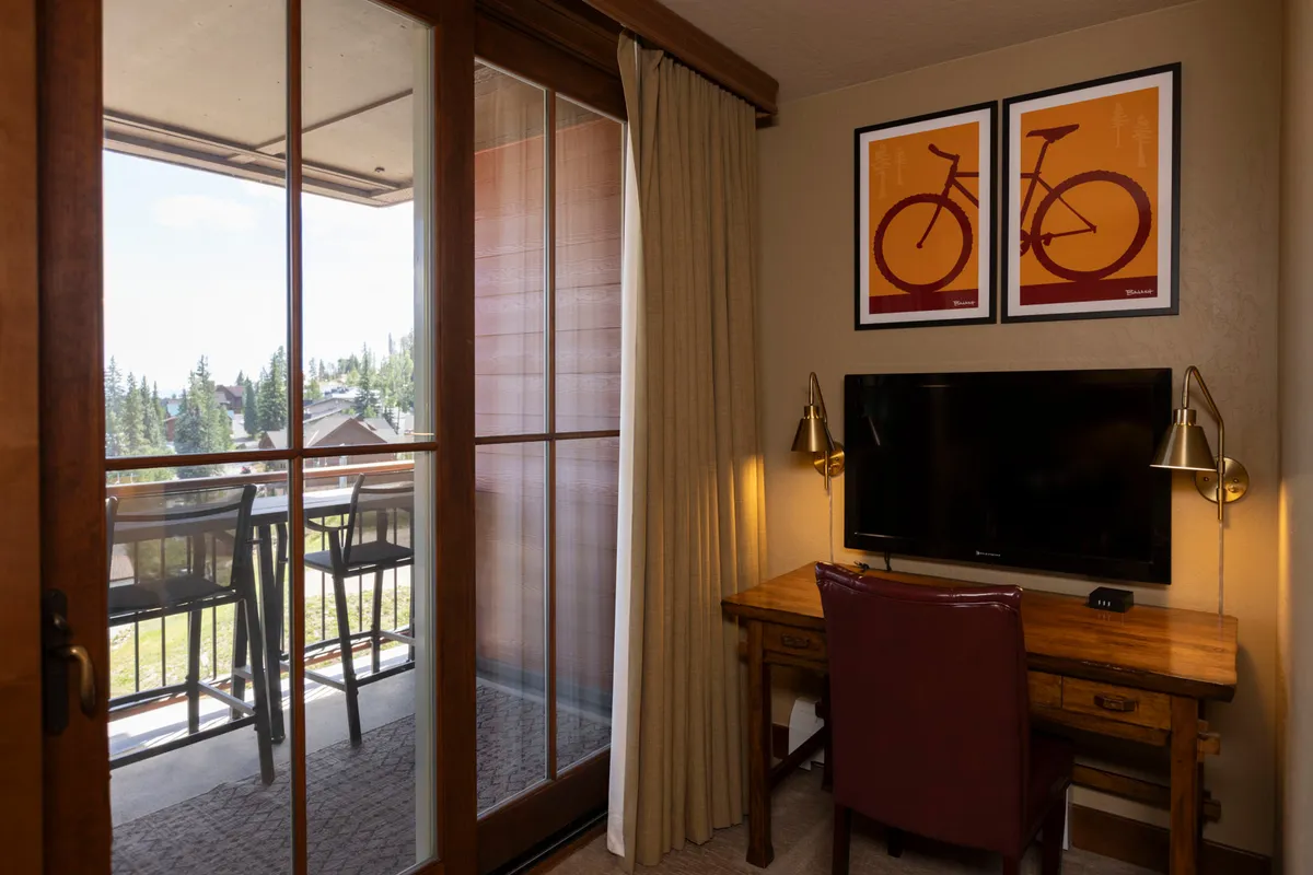 This interior shot features a comfortable workspace with a desk, chair, and television, complemented by creative bicycle artwork. Through the balcony doors, stunning mountain views are visible, blending productivity with artistic inspiration and a scenic backdrop.