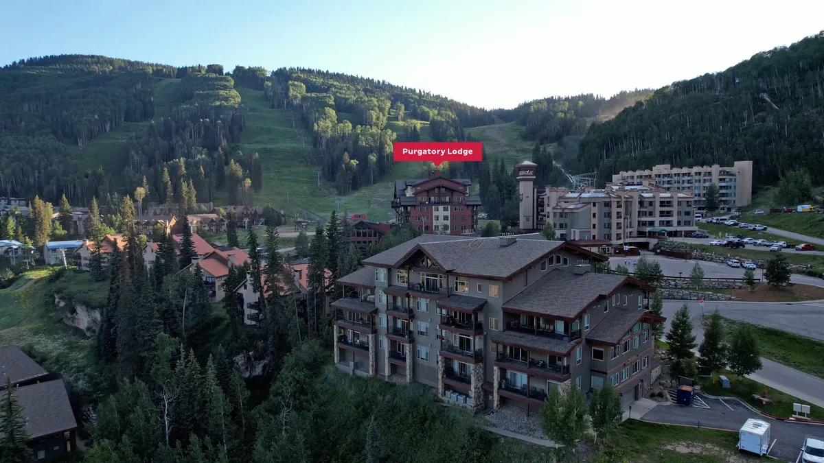 A wide-angle exterior view of Purgatory Lodge, surrounded by lush green trees and towering mountains, with visible outdoor spaces for gathering and recreation. The scene is serene yet adventurous, hinting at both relaxation and outdoor activities.