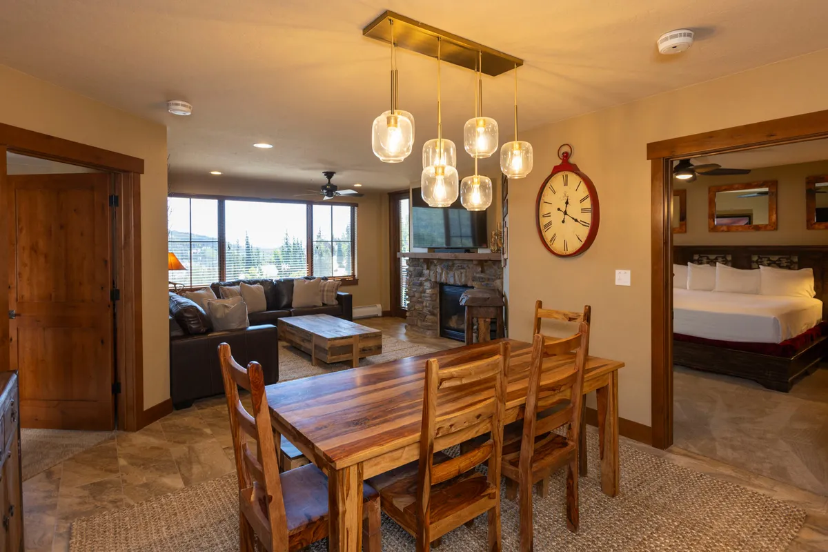 A sunlit, open-plan living and dining space featuring a spacious seating area, a glowing fireplace, modern pendant lighting, and large windows framing the scenic outdoors. The layout encourages conversation, relaxation, and shared experiences.