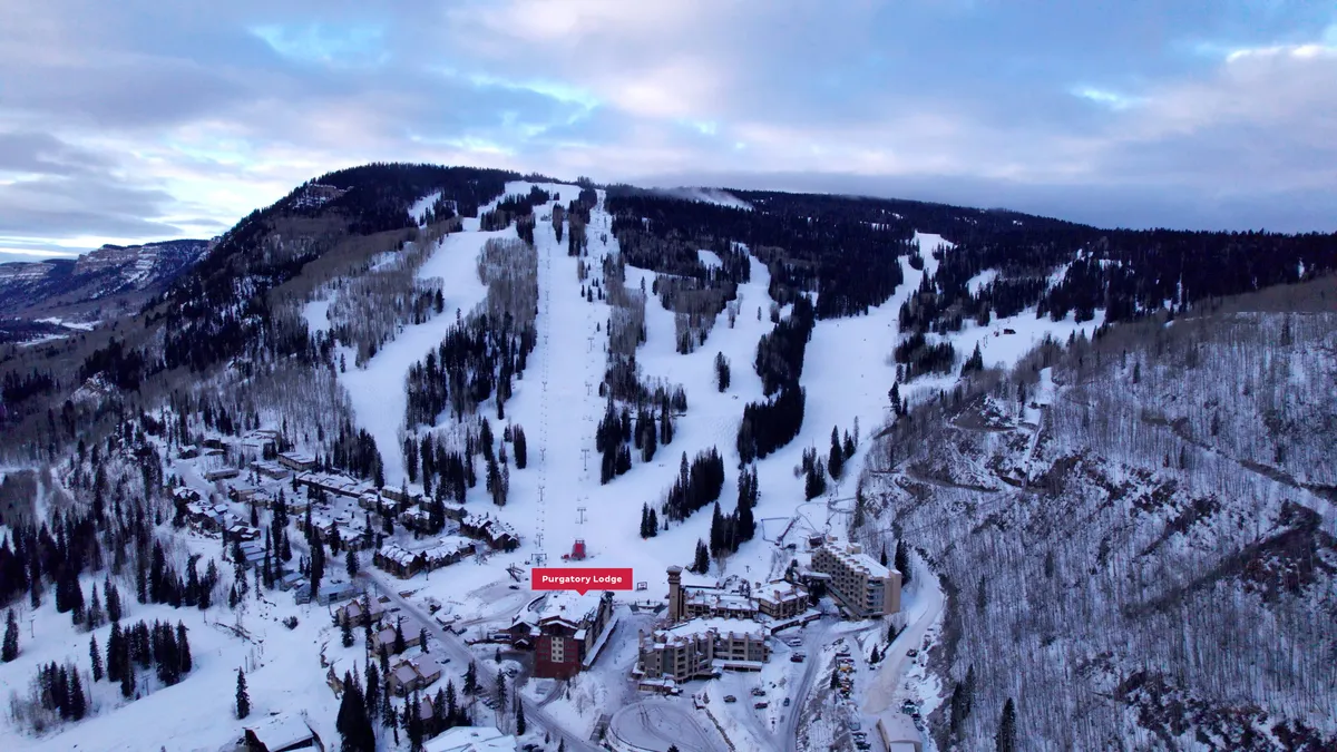This image features a sweeping exterior view of Purgatory Lodge set against the backdrop of snow-covered ski slopes, surrounding mountains, and a serene forest area. The scene captures both the lodge and easy slope access, inviting viewers to envision stepping outside and immediately starting their ski adventure.